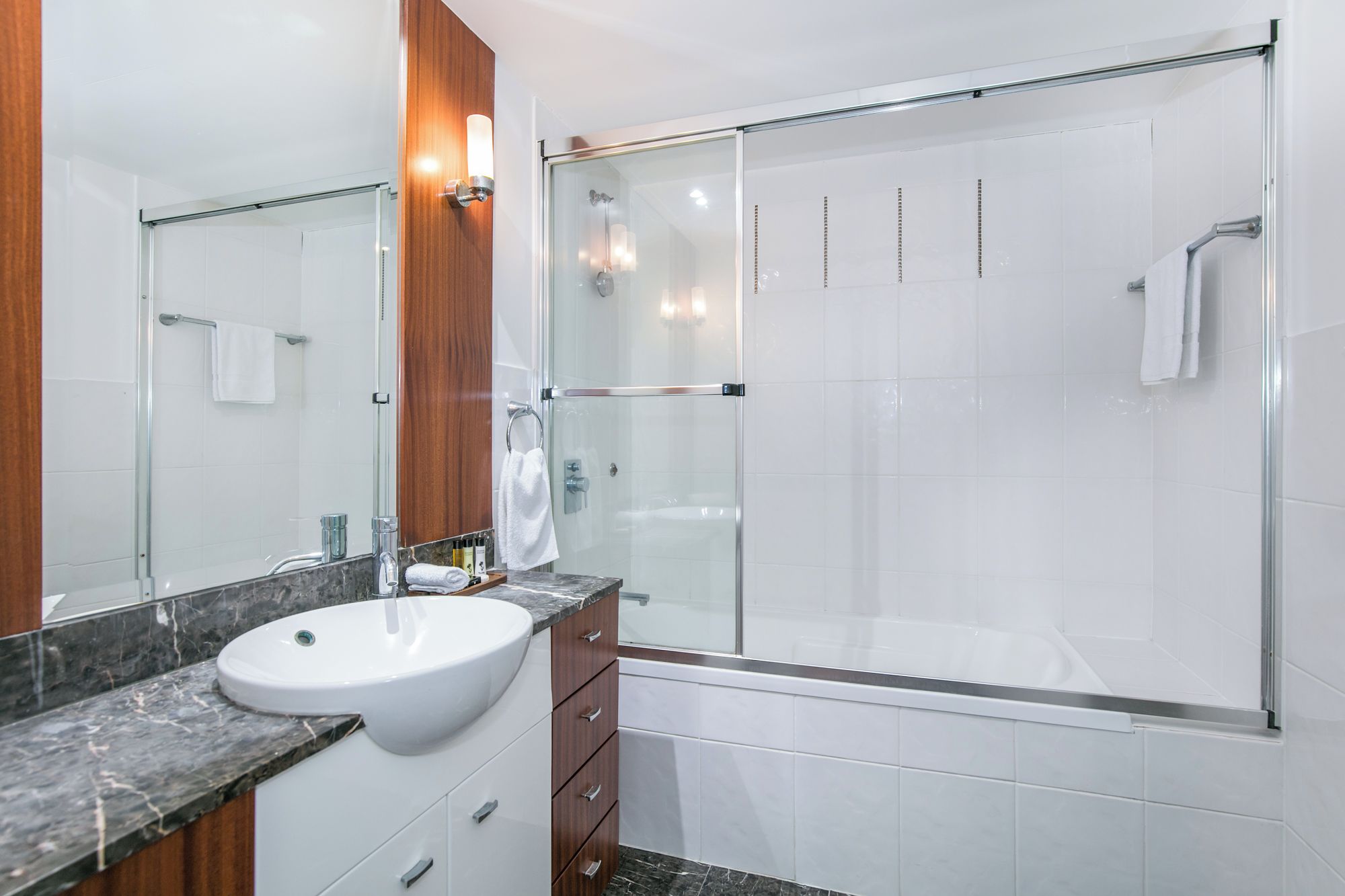 Un baño moderno con ducha envuelta en vidrio, lavamanos blanco y tocador de madera.