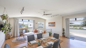 A spacious living area with a dining table set for four, a sofa, and a large window offering a view of the street. - Summer Sky - Oceanstays (Yamba)