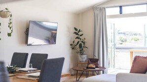 A modern living room with a dining area, a flat-screen TV, a potted plant, and a view through a large window. - Summer Sky - Oceanstays (Yamba)