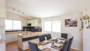 A modern kitchen with a central island, a dining area with a wooden table and chairs, and a large window providing natural light. - Summer Sky - Oceanstays (Yamba)