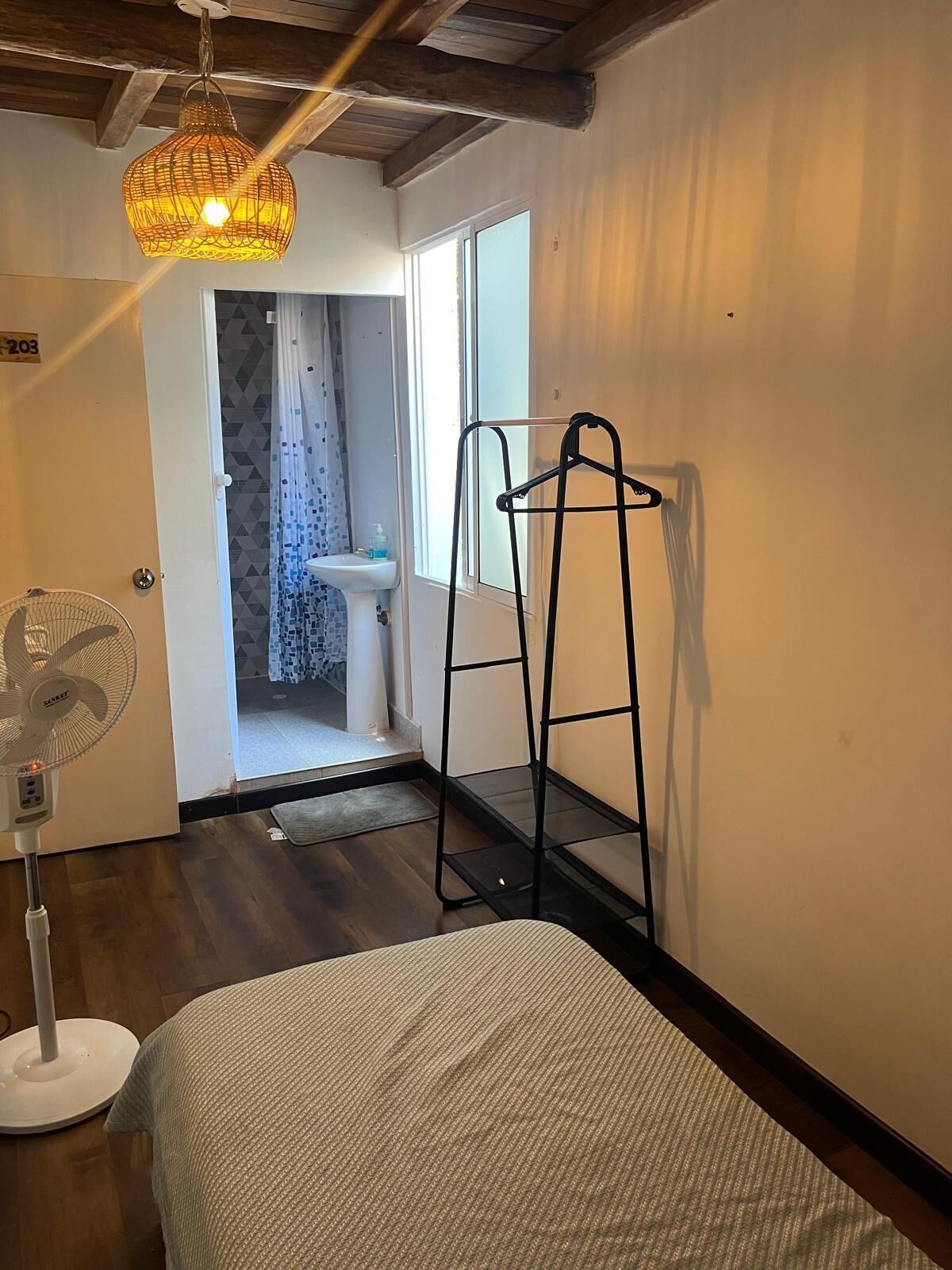 A room with a bed, a standing fan, a black metal rack, and a hanging light fixture.