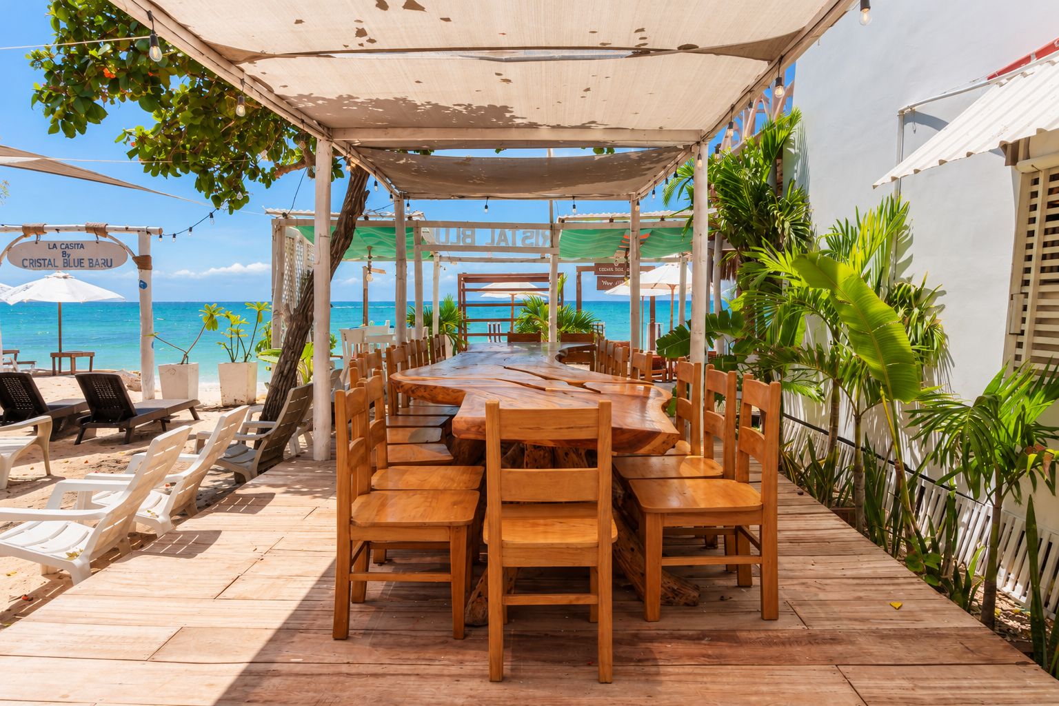 An outdoor dining area with a long wooden table and chairs, surrounded by a shaded canopy, overlooking a beach with a clear view of the ocean.