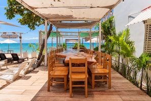 Un espace de restauration en plein air avec une longue table et des chaises en bois, entouré d’un auvent ombragé, offrant une vue dégagée sur une plage et l’océan.