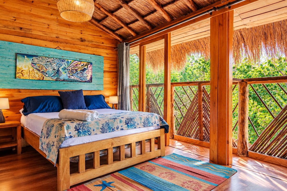 A wooden room with a bed, a painting, and a view of greenery.