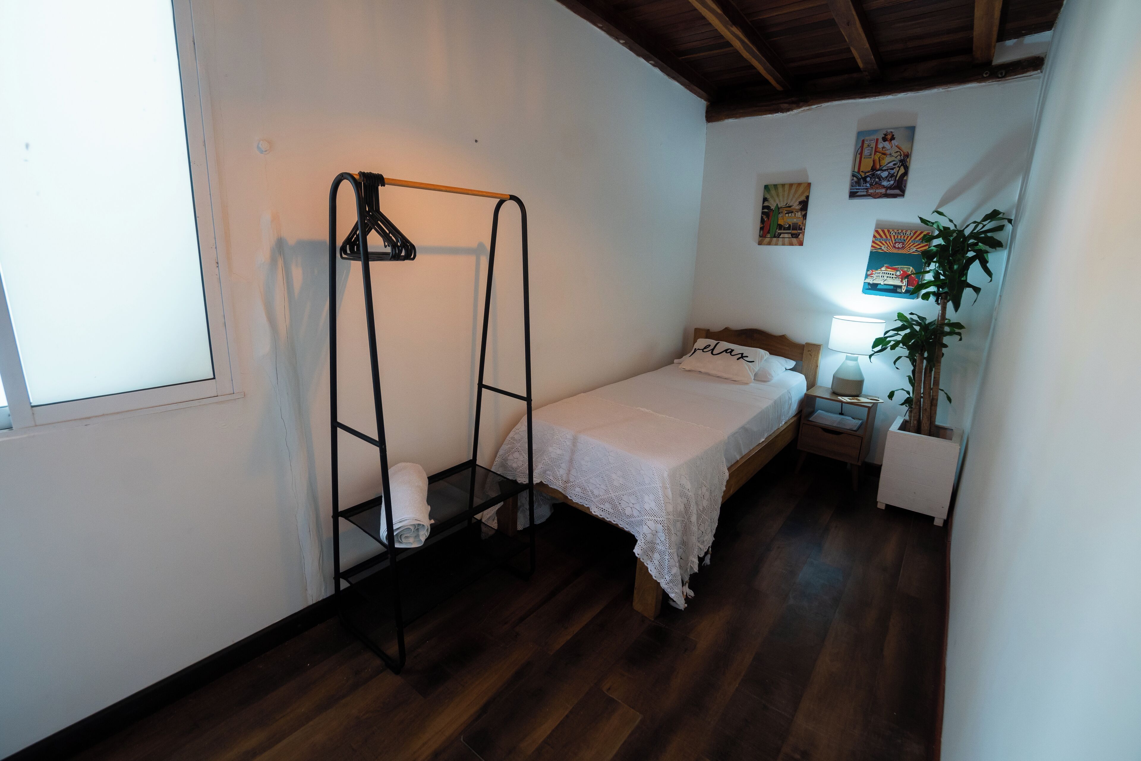 A small, neatly arranged bedroom with a single bed, a nightstand, a bedside lamp, a hanging rack, and a window.