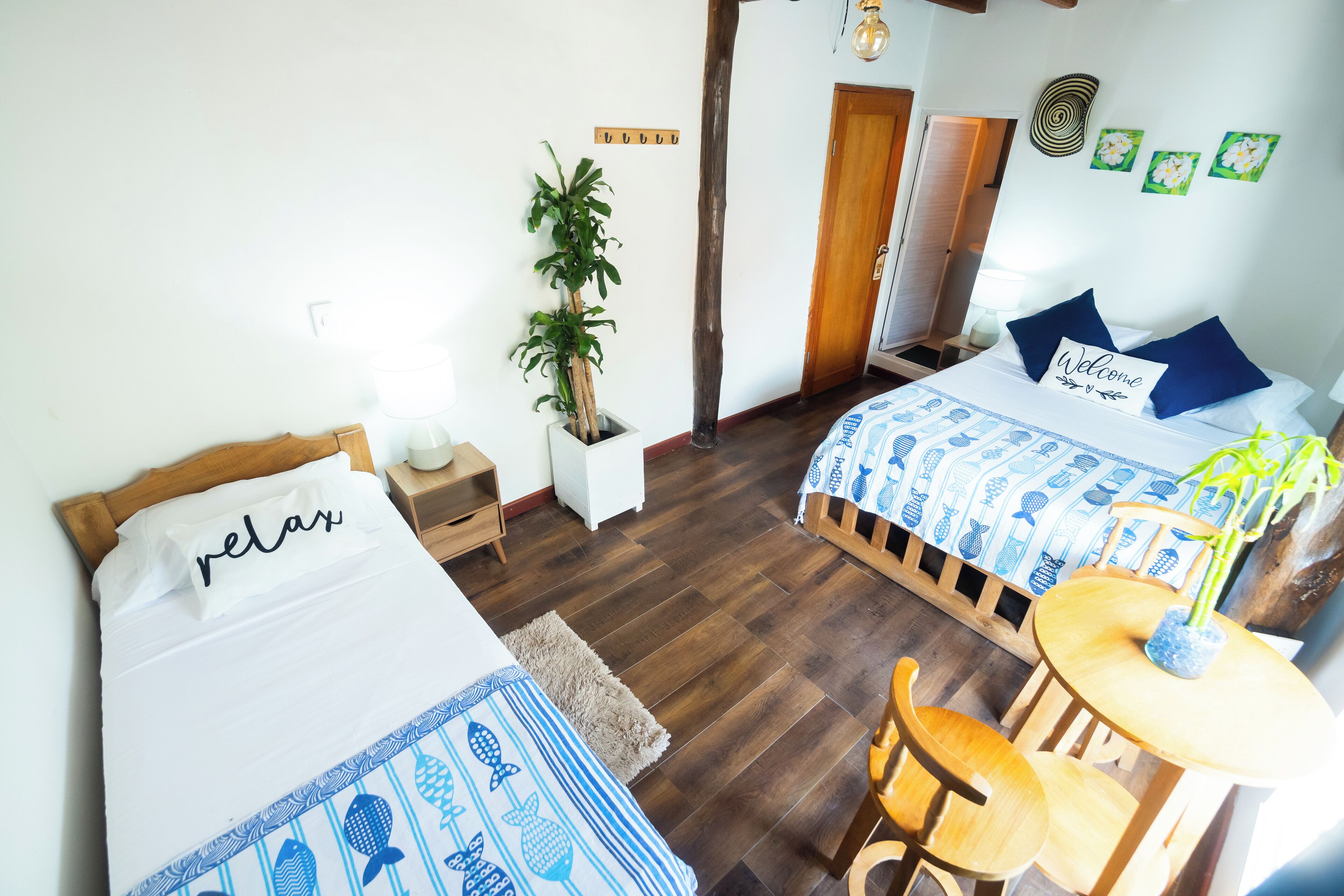 A hotel room with two beds, a wooden floor, a small table with a chair, and a potted plant.