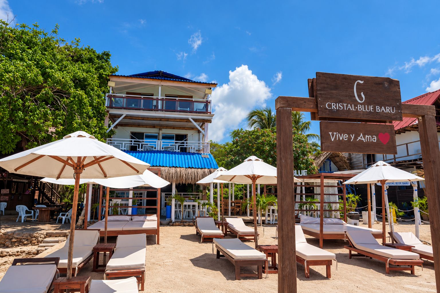 Un resort frente a la playa con sillas de descanso y sombrillas, un cartel que dice 'Cristal Blue Baru', y un edificio con techo azul.