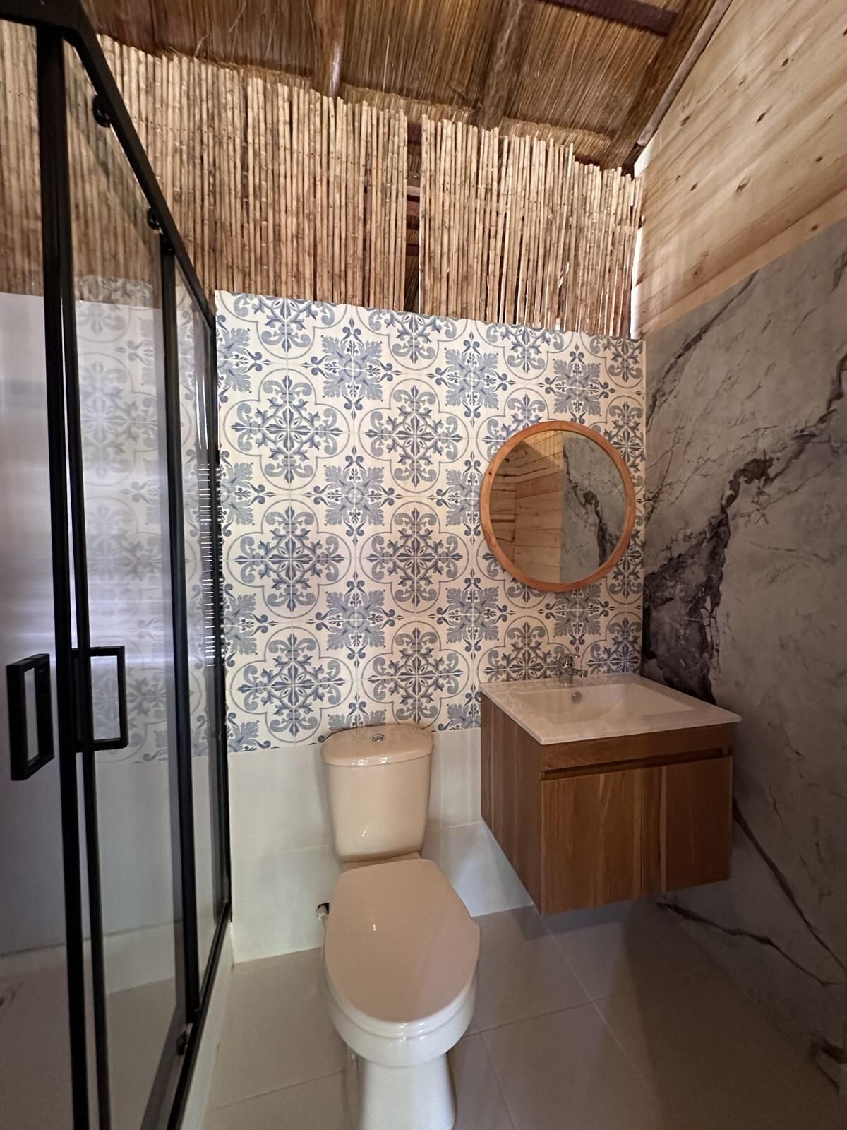 A bathroom with a toilet, a wooden cabinet, a glass shower enclosure, and a wall with patterned tiles.