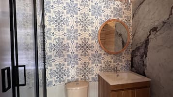 A bathroom with a toilet, a wooden cabinet, a glass shower enclosure, and a wall with patterned tiles.