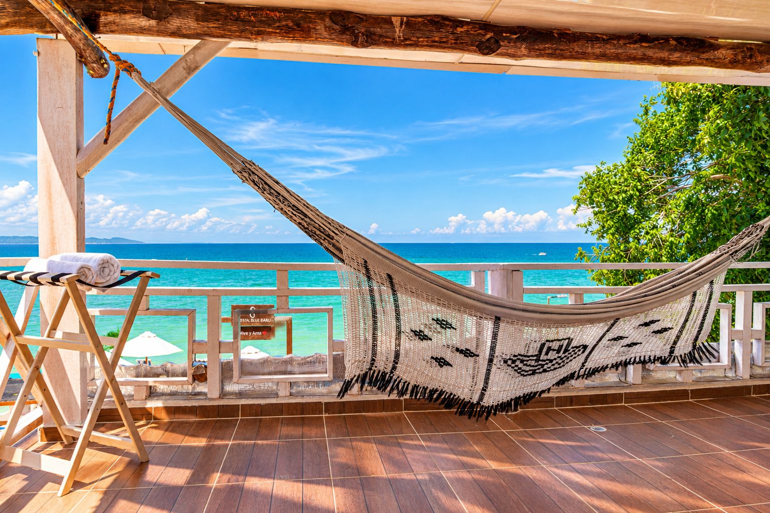 A hammock on a wooden deck overlooking a turquoise sea, with a wooden railing and a sign indicating a beach view.