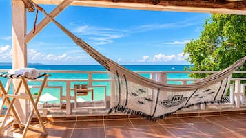 A hammock on a wooden deck overlooking a turquoise sea, with a wooden railing and a sign indicating a beach view.