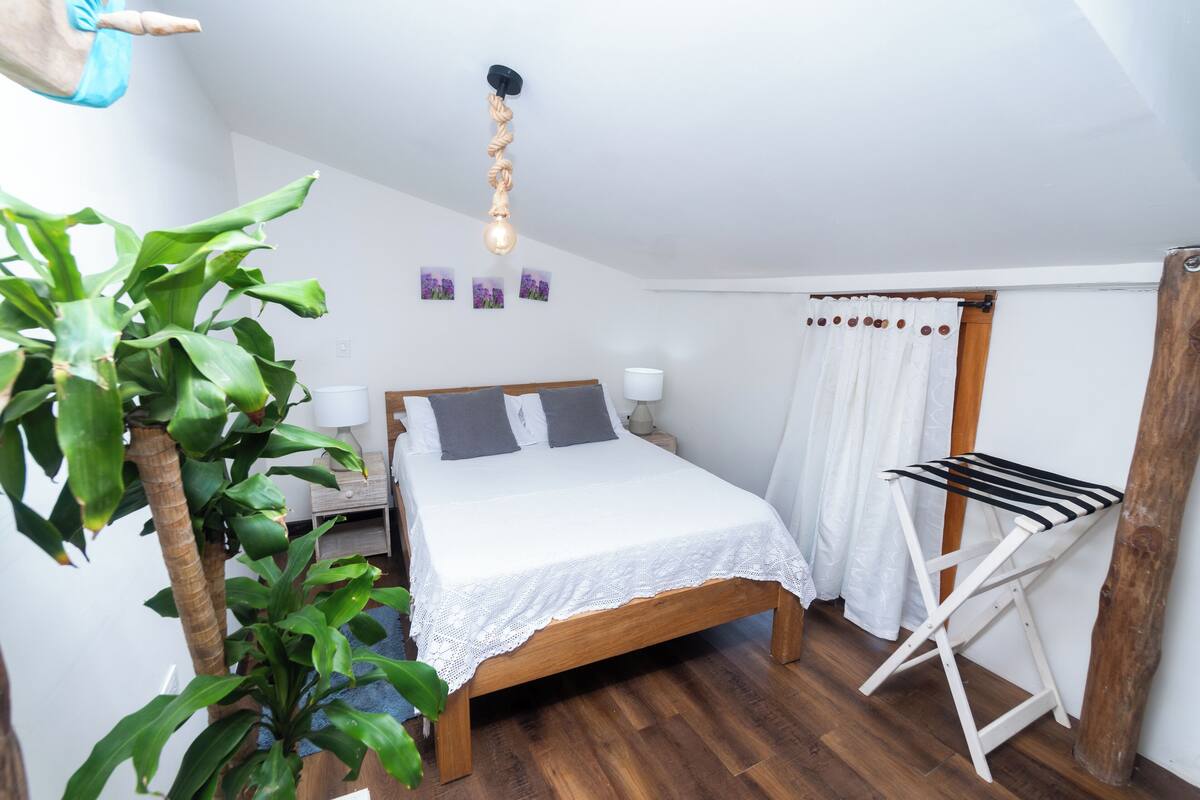 A bedroom with a wooden bed, white bedding, a hanging light fixture, a plant, and a drying rack.