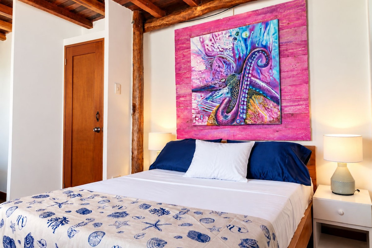 Ein Schlafzimmer mit einem Bett, einem Nachttisch, einer Lampe und einem großen, abstrakten Gemälde an der Wand.