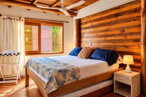 Une chambre à coucher avec une tête de lit en bois, un lit avec une literie bleue et blanche, une table de chevet avec une lampe, et une fenêtre donnant sur un paysage verdoyant.
