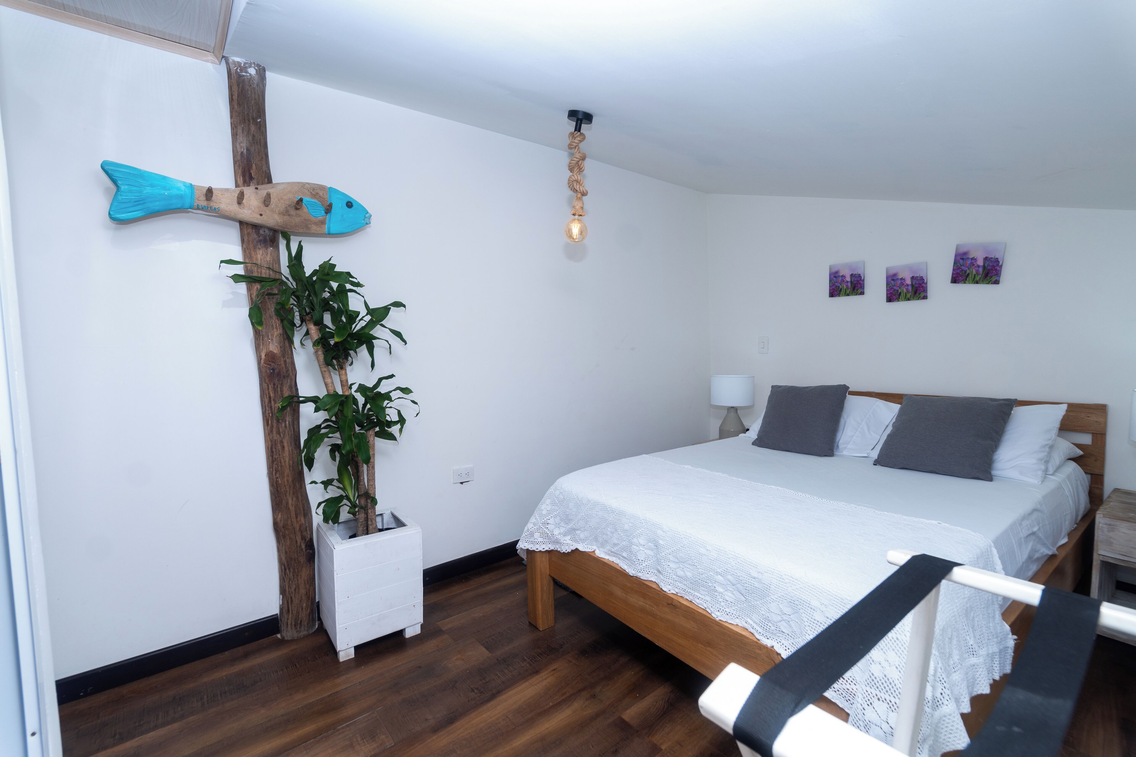 A bedroom with a wooden bed, white bedding, a wall-mounted fish decoration, a potted plant, and a ceiling light fixture.