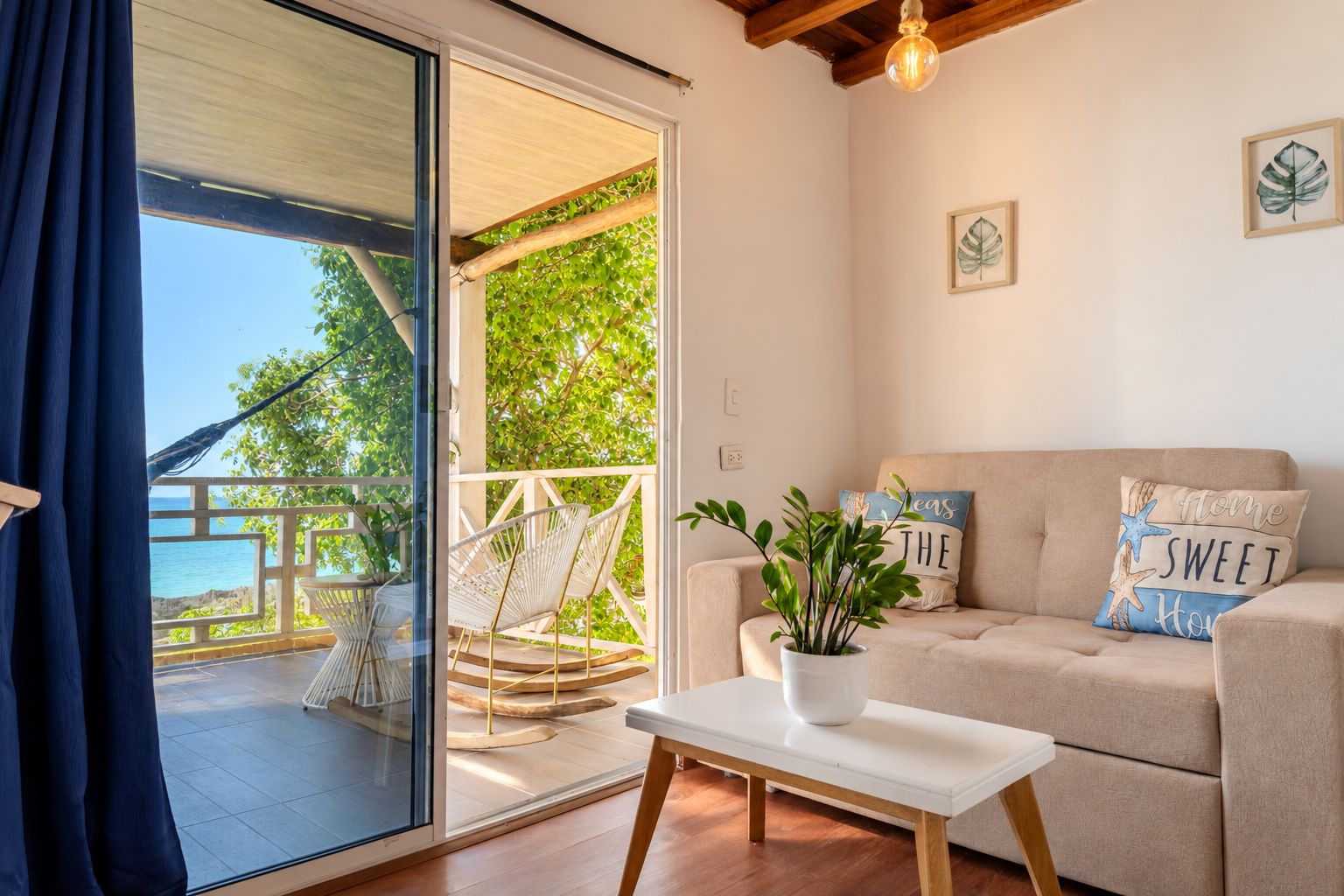 Una habitación con puerta de vidrio corredera que da a un balcón con vistas al mar. Hay un sofá con almohadones decorativos y una mesita con una planta.