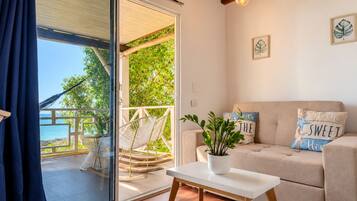 Una habitación con puerta de vidrio corredera que da a un balcón con vistas al mar. Hay un sofá con almohadones decorativos y una mesita con una planta.