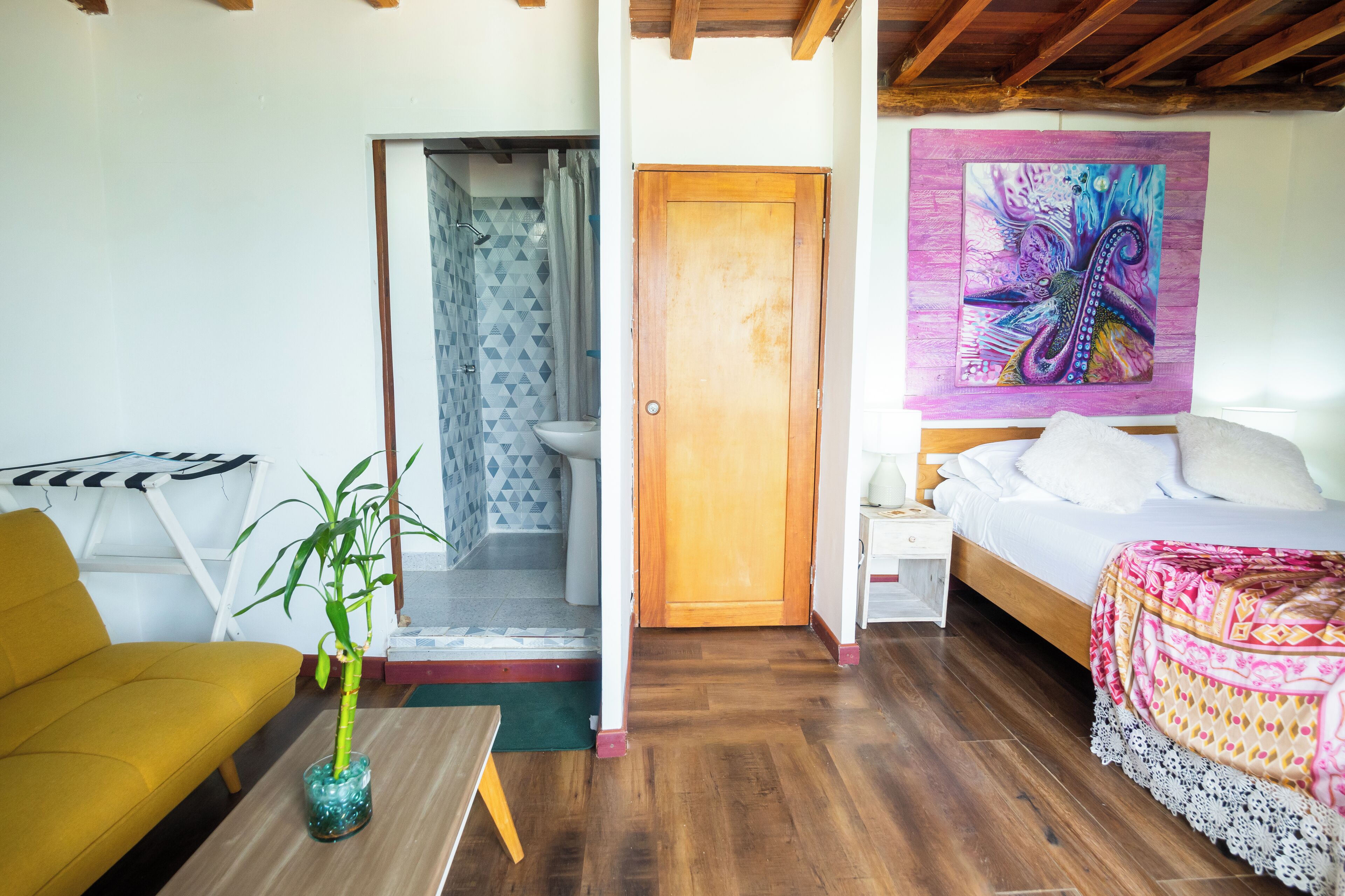 Una habitación con una cama, un sofá, suelo de madera, baño con ducha y un cuadro de colores en la pared.