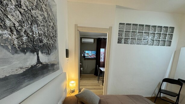 Ein Schlafzimmer mit Bett, Nachttisch, Lampe, einem Bild, einem Fernseher im Hintergrund und einem Fenster mit Jalousien.