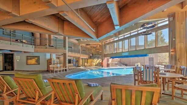 A spacious indoor pool area with wooden lounge chairs and a view of the outside. - Cozy Ski-In/Out Studio In Building 4 | Kitchen | Hot Tubs, Dog Friendly (Breckenridge)