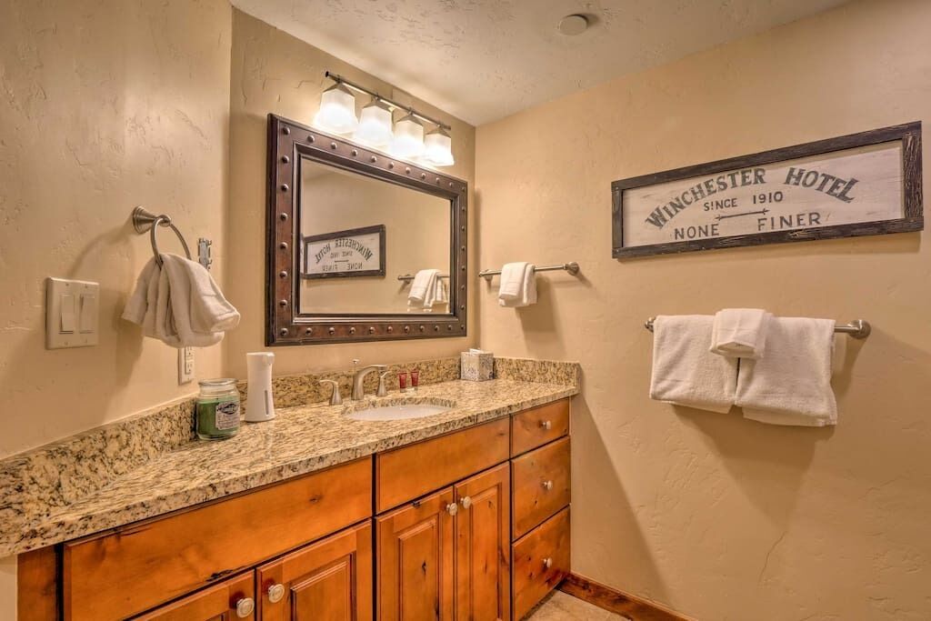 A bathroom with a granite countertop, wooden cabinets, a large mirror with lights, and a sign that reads "Winchester Hotel Since 1910 None Finer."