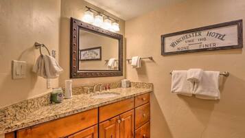 A bathroom with a granite countertop, wooden cabinets, a large mirror with lights, and a sign that reads "Winchester Hotel Since 1910 None Finer."