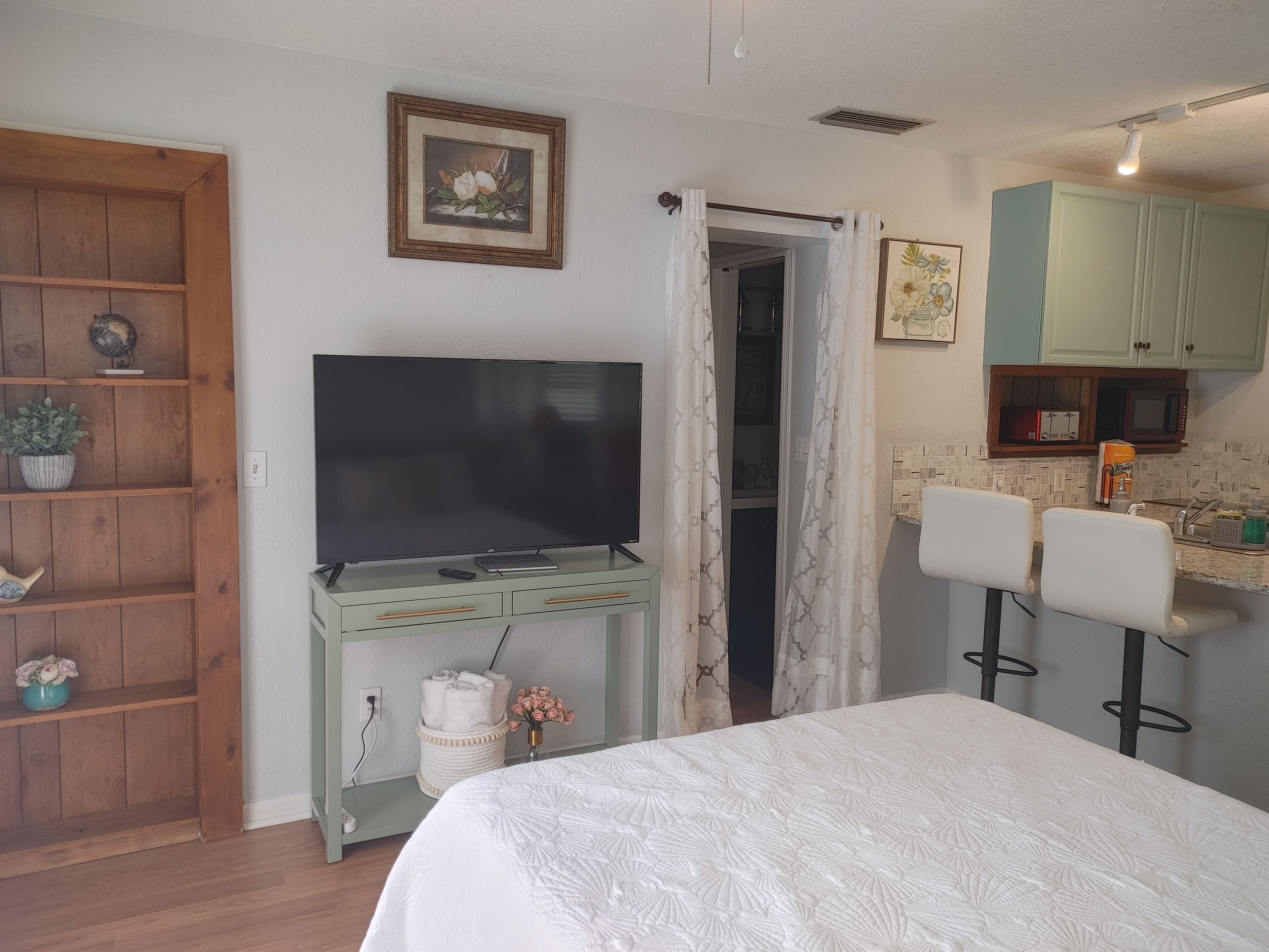 A bedroom with a bed, a TV on a wooden stand, a bathroom with a shower, and a kitchenette with a microwave.