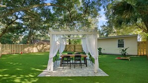Eine Terrasse mit Tisch und Stühlen unter einer weißen Markise, umgeben von einem gepflegten Rasen und einem Holzzaun.