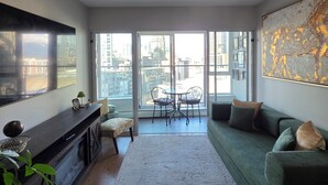A modern living room with a large window, a green sofa, a dining table with chairs, and a flat-screen TV on a wooden console. - Charming Dwelling Downtown Vancouver (Vancouver)