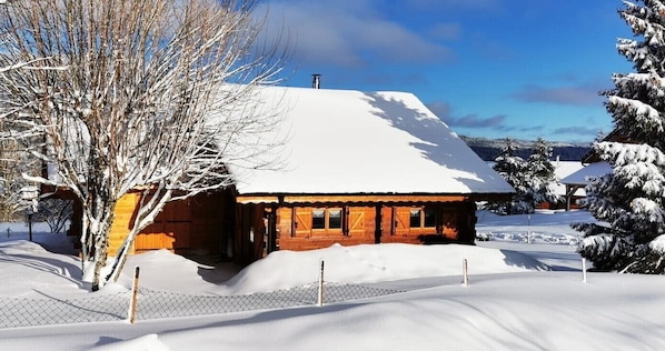 Une cabane en bois dont le toit est recouvert de neige, entourée d’arbres également couverts de neige, sous un ciel dégagé.