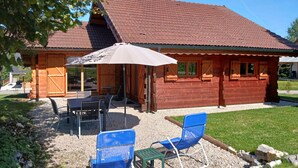 A wooden cabin with a tiled roof, a covered patio with a table and chairs, and a gravel area with two blue chairs. - Les 7 Coeurs, cosy chalet in Val Foncine (Foncine-le-Haut)
