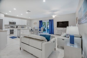 A modern, well-lit living area with a sofa, a coffee table, a TV, and a dining area with chairs and a table.