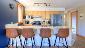 Une cuisine moderne dotée d’un îlot central, de tabourets de bar, de meubles en bois, d’appareils électroménagers en acier inoxydable et d’une fenêtre avec des stores.