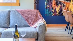 Un salon avec un canapé gris, une table basse en bois sur laquelle se trouvent une bouteille de vin et un verre, une tapisserie colorée accrochée au mur et un fauteuil.