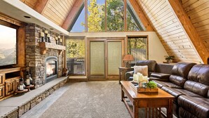 A cozy living room with a stone fireplace, leather sofa, and a large window with a view of trees. - Amazing A Frame w/ Gorgeous Views of Bear Mountain + Spa (Big Bear Lake)