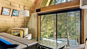 A cozy cabin interior with wooden walls, a large window with blinds, a ceiling lamp, and a small table with a grill. - Amazing A Frame w/ Gorgeous Views of Bear Mountain + Spa (Big Bear Lake)