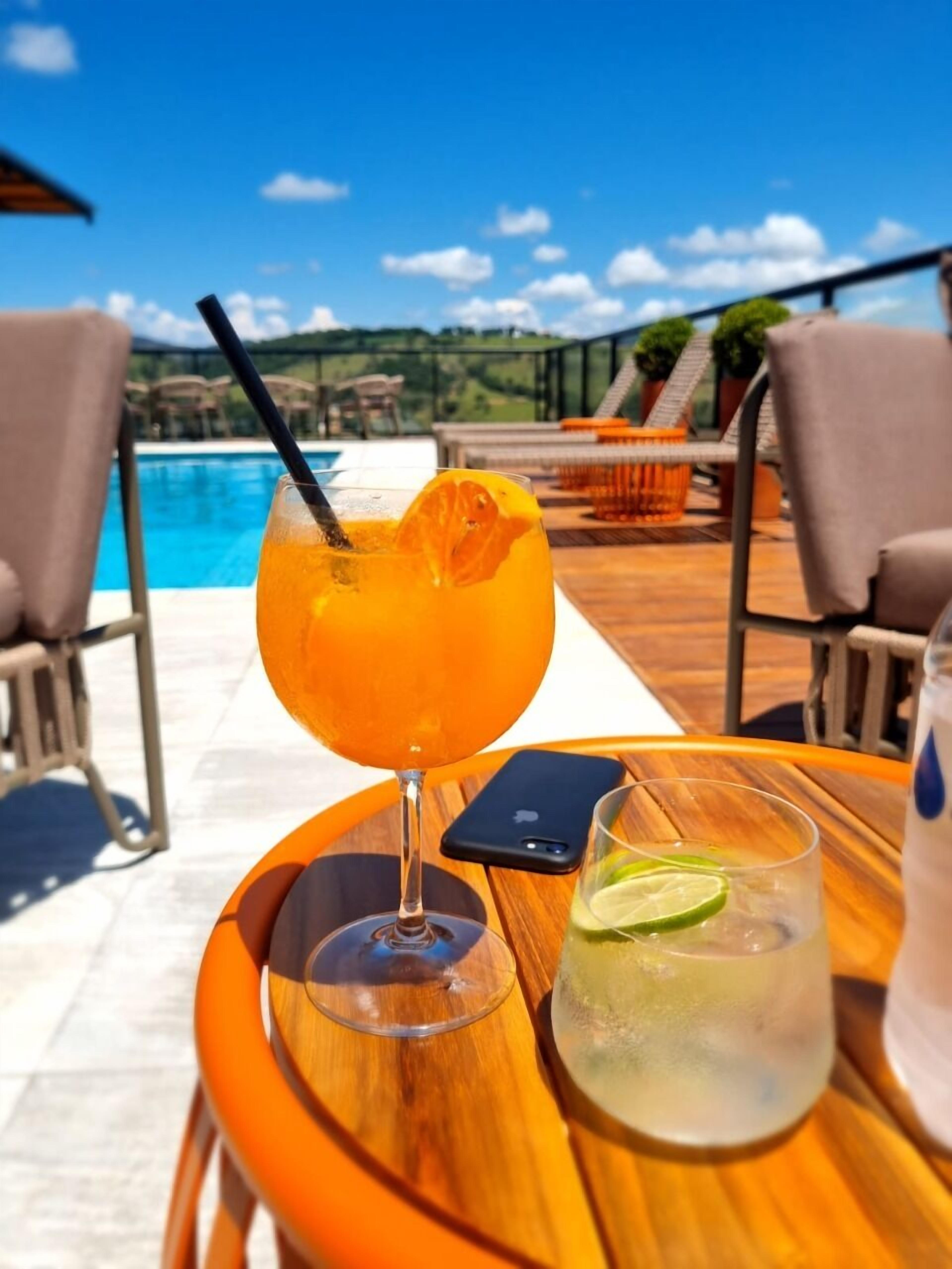 A wooden table with a glass of orange cocktail and a glass of lime water, a smartphone, and a pool in the background.