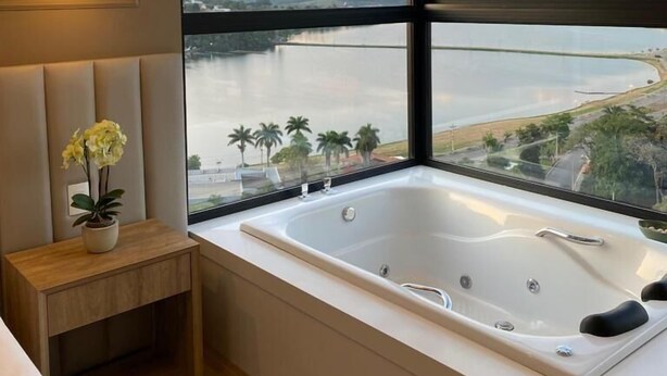 A modern bathroom with a large bathtub, a small wooden side table with a potted plant, and a view of a lake and mountains through large windows. - Vista do Lago Hotel (Capitólio)