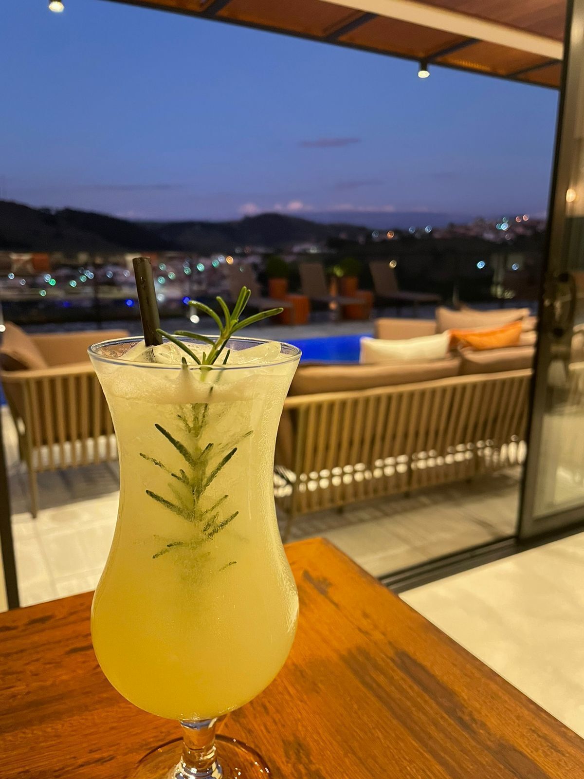 Ett cocktailglas med sugrör och en rosmarinkvist som garnering, placerat på ett träbord med utsikt över en pool och ett loungeområde med utomhusmöbler.