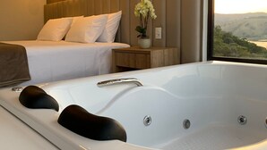 A modern bedroom with a large bed, a built-in bathtub, and a view of a landscape through a large window. - Vista do Lago Hotel (Capitólio)