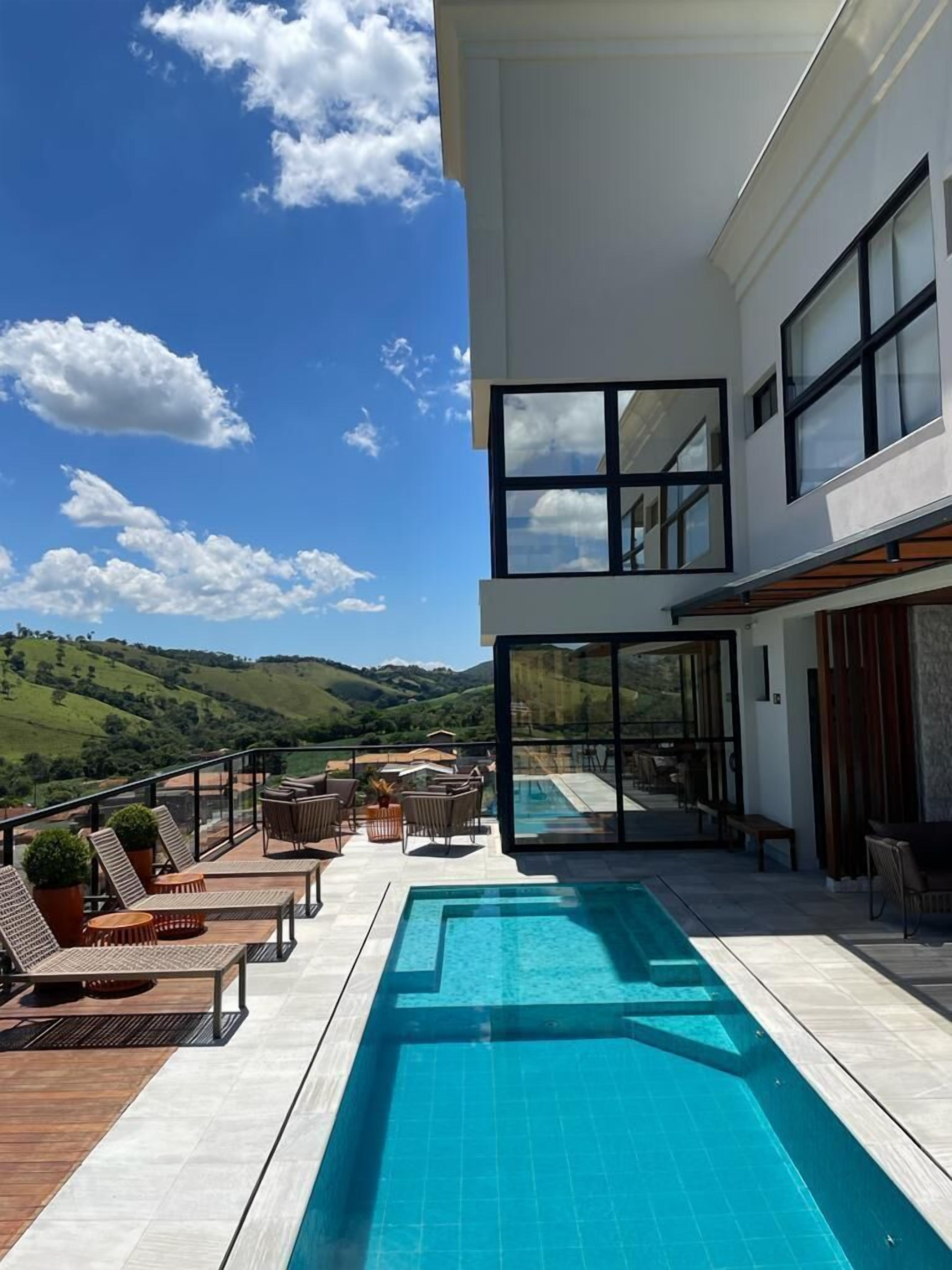 A modern two-story building with a swimming pool, outdoor seating, and a view of green hills.