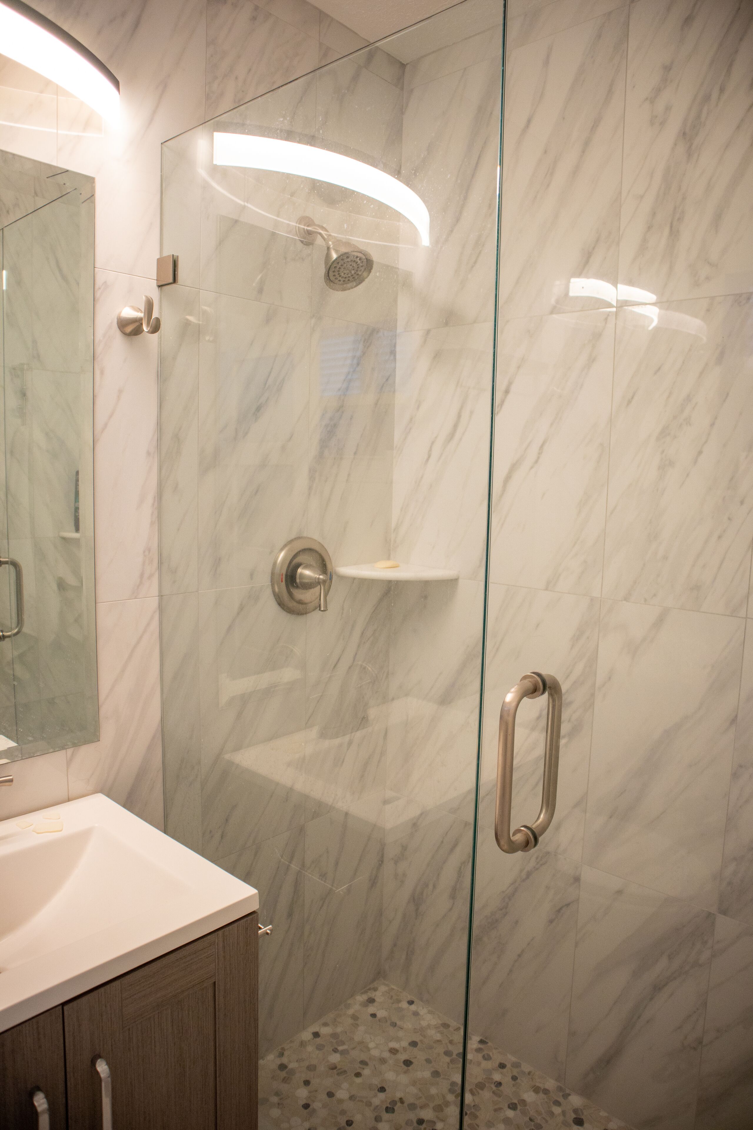 A modern bathroom with a glass-enclosed shower, a marble-tiled wall, and a white sink with a wooden cabinet.