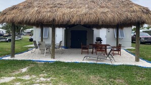 Une construction à toit de chaume, avec une table et des chaises, entourée d’une zone sablonneuse et d’une pelouse verdoyante.