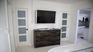 Une chambre avec un lit, une commode, une télévision et, au fond, une cuisine.