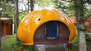 Una estructura esférica con una puerta azul y bolas de colores por dentro, rodeada de árboles y un edificio rojo al fondo.