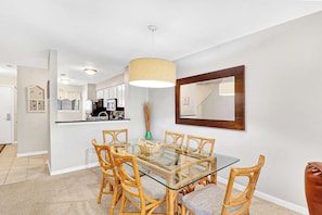 A dining area with a glass table and wicker chairs, a kitchen in the background, and a large mirror on the wall. - Walk to the Beach Springwood Villa Retreat (Hilton Head Island)
