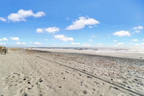 Une vaste plage de sable où l’on peut voir des empreintes de pas qui mènent vers l’océan, sous un ciel bleu dégagé parsemé de quelques nuages.