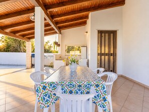 Patio esterno con tavolo apparecchiato per mangiare, circondato da sedie bianche, sotto un tetto in legno.