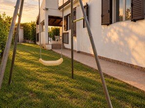 A residential area with a swing set, a paved walkway, and a building with a balcony. - MarinaBlue Haven (Santa Maria del Focallo)
