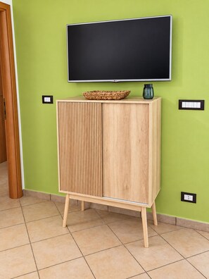 A room with a green wall, a wooden cabinet with a flat-screen TV mounted above it, and a tiled floor. - MarinaBlue Haven (Santa Maria del Focallo)